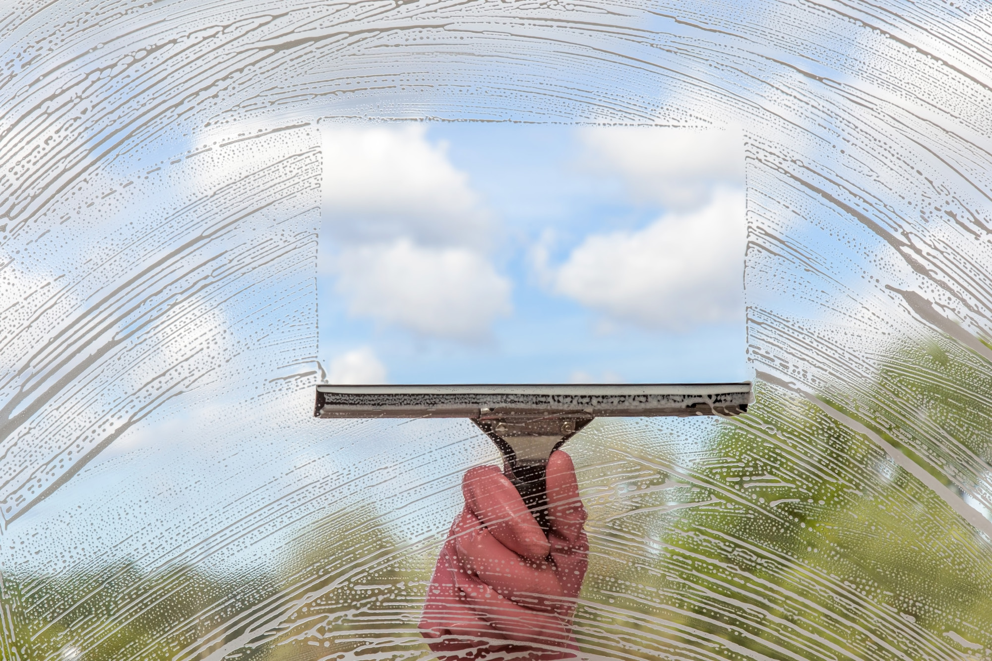 Hand in protective glove washing and cleaning window with professionally squeegee on background of cloudy sky. Summer windows cleaning. Maid cleans window.